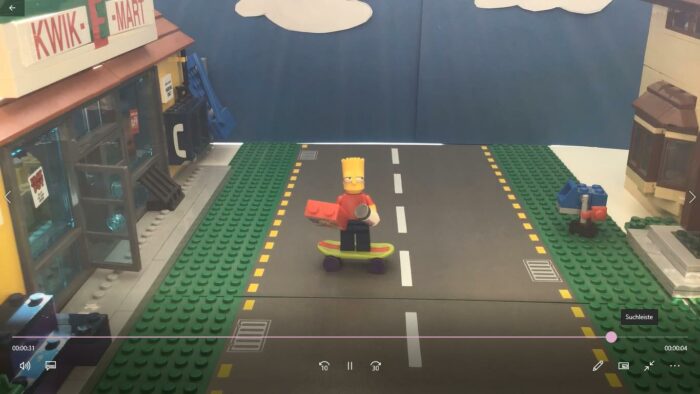Lego-Figur auf Skateboard vor einem Kwik-E-Mart-Gebäude auf einer Straße mit grünen Rändern.
